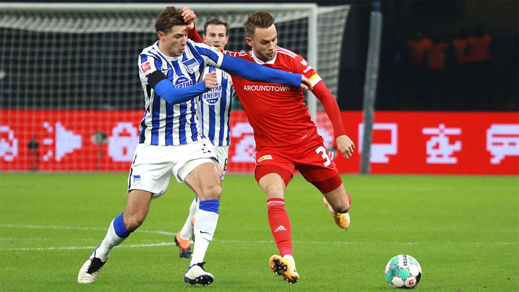 Zwei Fußballspieler kämpfen um den Ball während eines Spiels. Einer trägt ein blau-weißes Trikot, der andere rot.