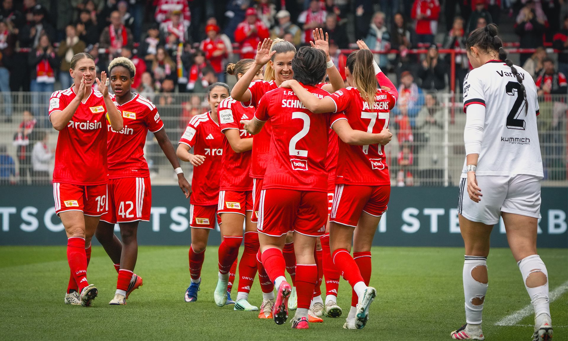 Eine Gruppe von Fußballspielerinnen in roten Trikots feiert ein Tor, während eine gegnerische Spielerin im Hintergrund steht.