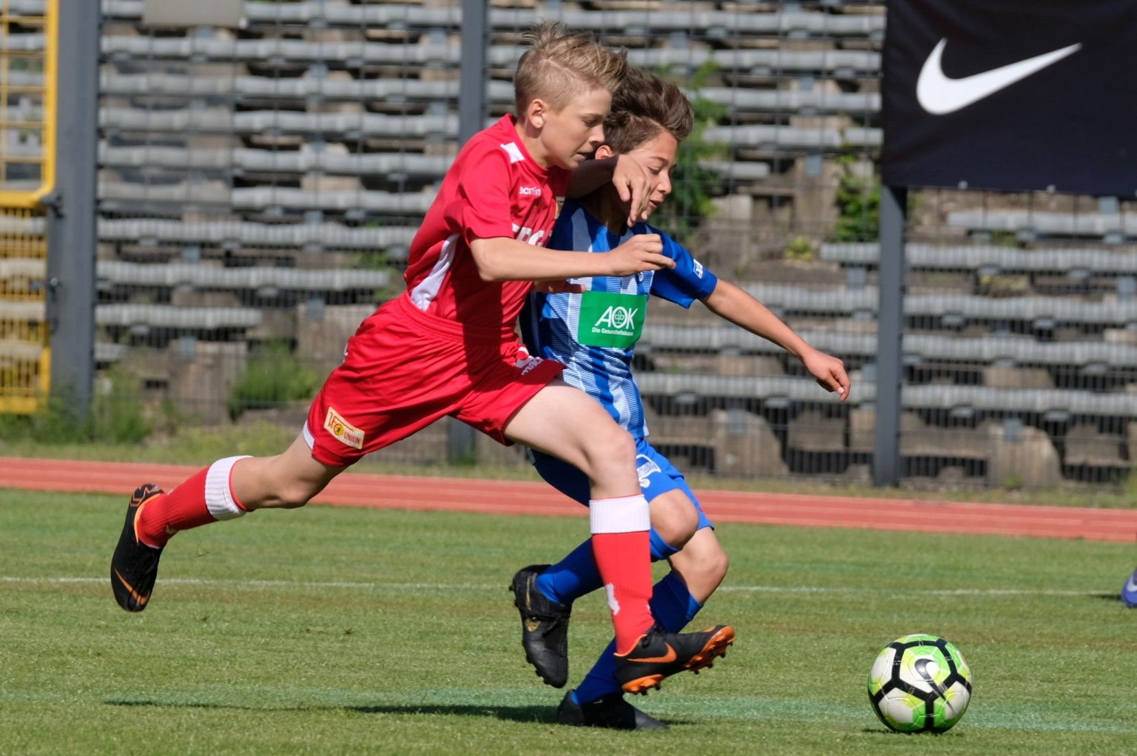 Zwei junge Fußballspieler im Wettkampf um den Ball auf einem Sportplatz. Einer trägt ein rotes, der andere ein blaues Trikot.