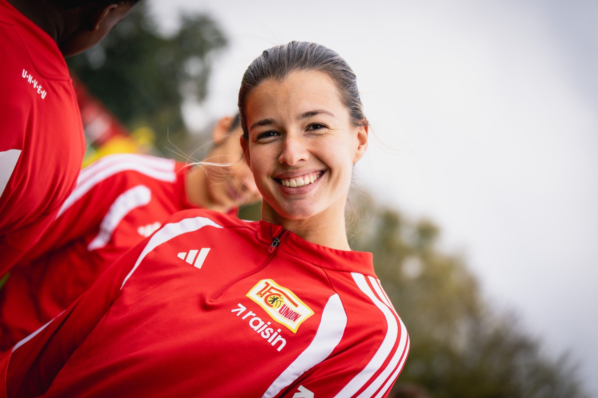 Eine lächelnde Frau in roter Sportbekleidung mit dem Logo eines Fußballvereins im Hintergrund.
