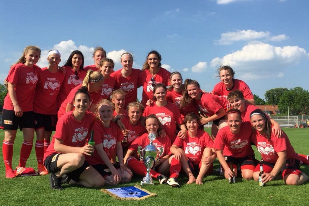 Eine Gruppe von Frauen in roten T-Shirts steht auf einem Fußballfeld und posiert mit einem Pokal und Urkunde.
