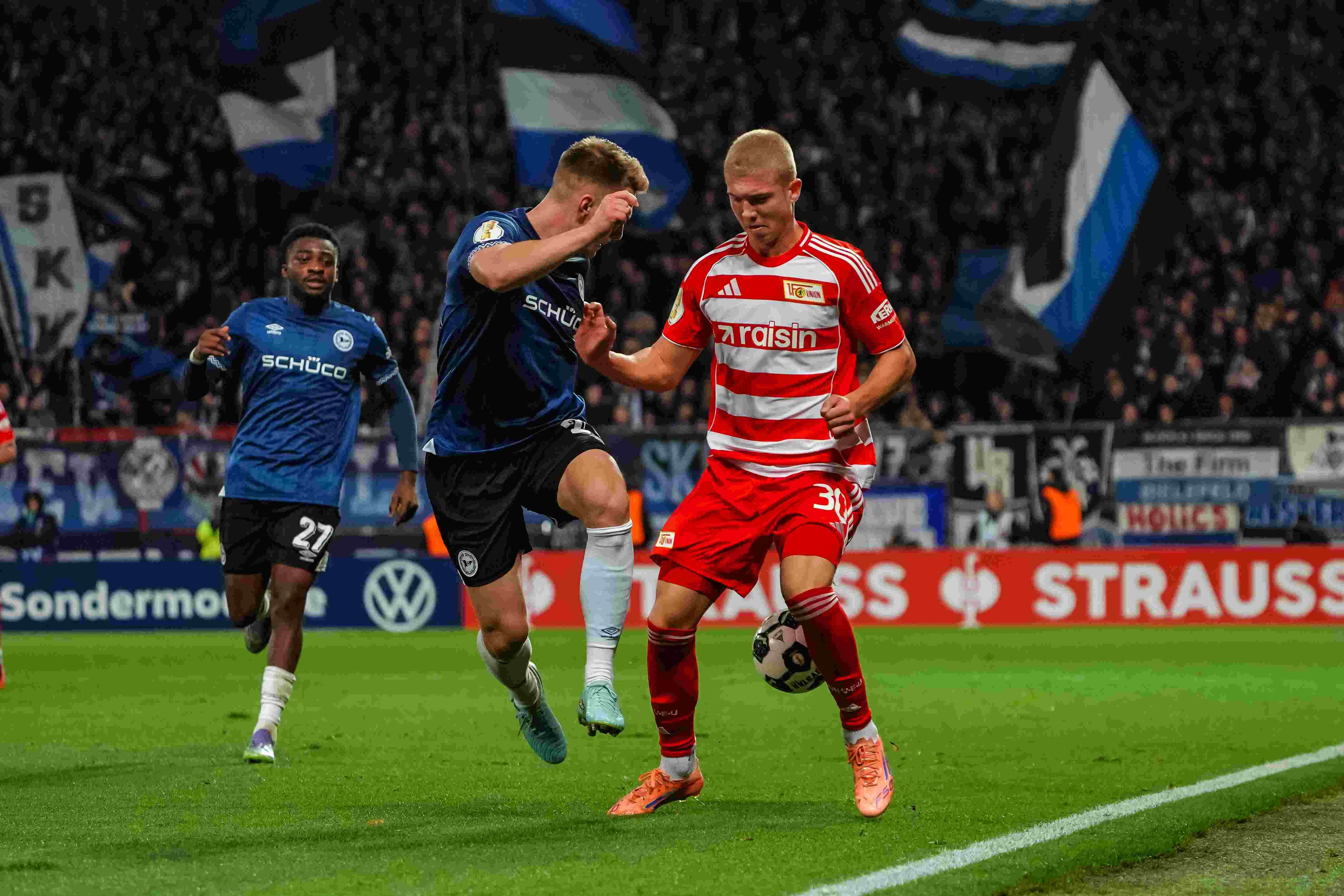 Ein Fußballspieler mit blauem Trikot springt über einen Spieler mit rotem Trikot. Im Hintergrund sind die Fans und Fahnen.