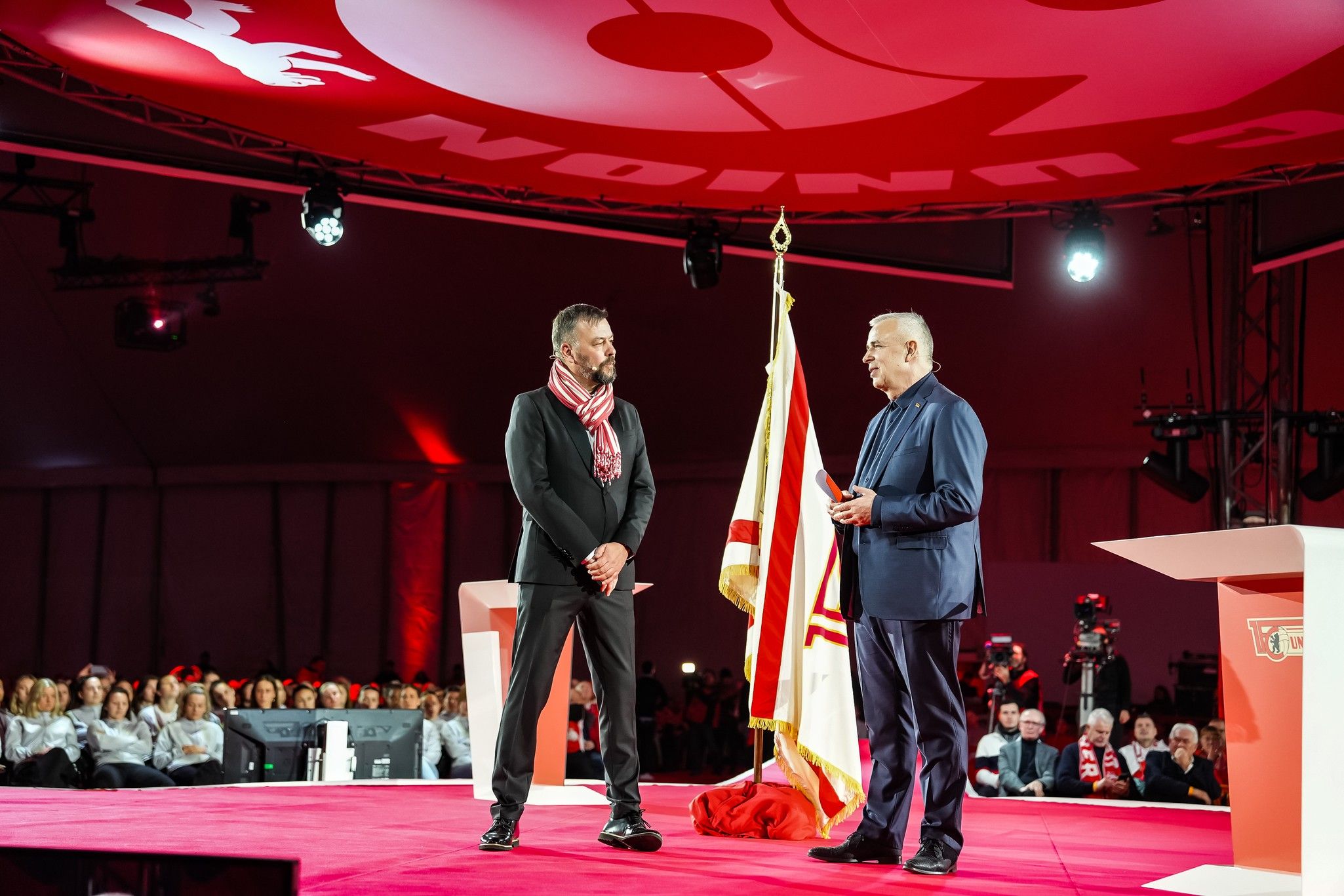 Zwei Männer stehen auf einer Bühne vor einem Publikum, einer hält eine Flagge und spricht, während der andere zuhört.