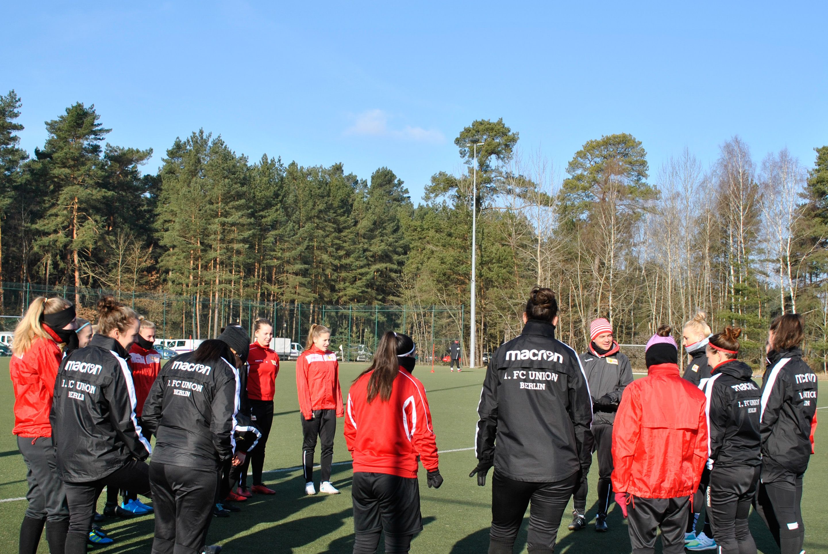 Die Spielerinnen einer Fußballmannschaft stehen in einem Kreis auf einem Platz, umgeben von Bäumen, während sie sich unterhalten.
