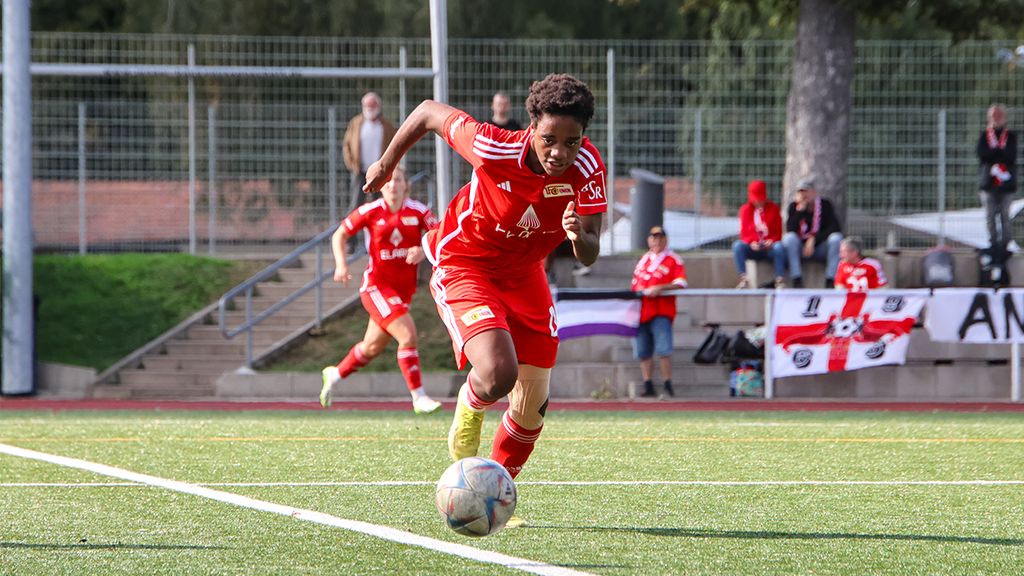Spielerin in rotem Trikot sprintet über das Fußballfeld, während Zuschauer im Hintergrund zuschauen.
