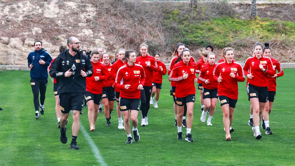 A group of female soccer players in red tracksuits jogs on a grass field under the guidance of a coach.