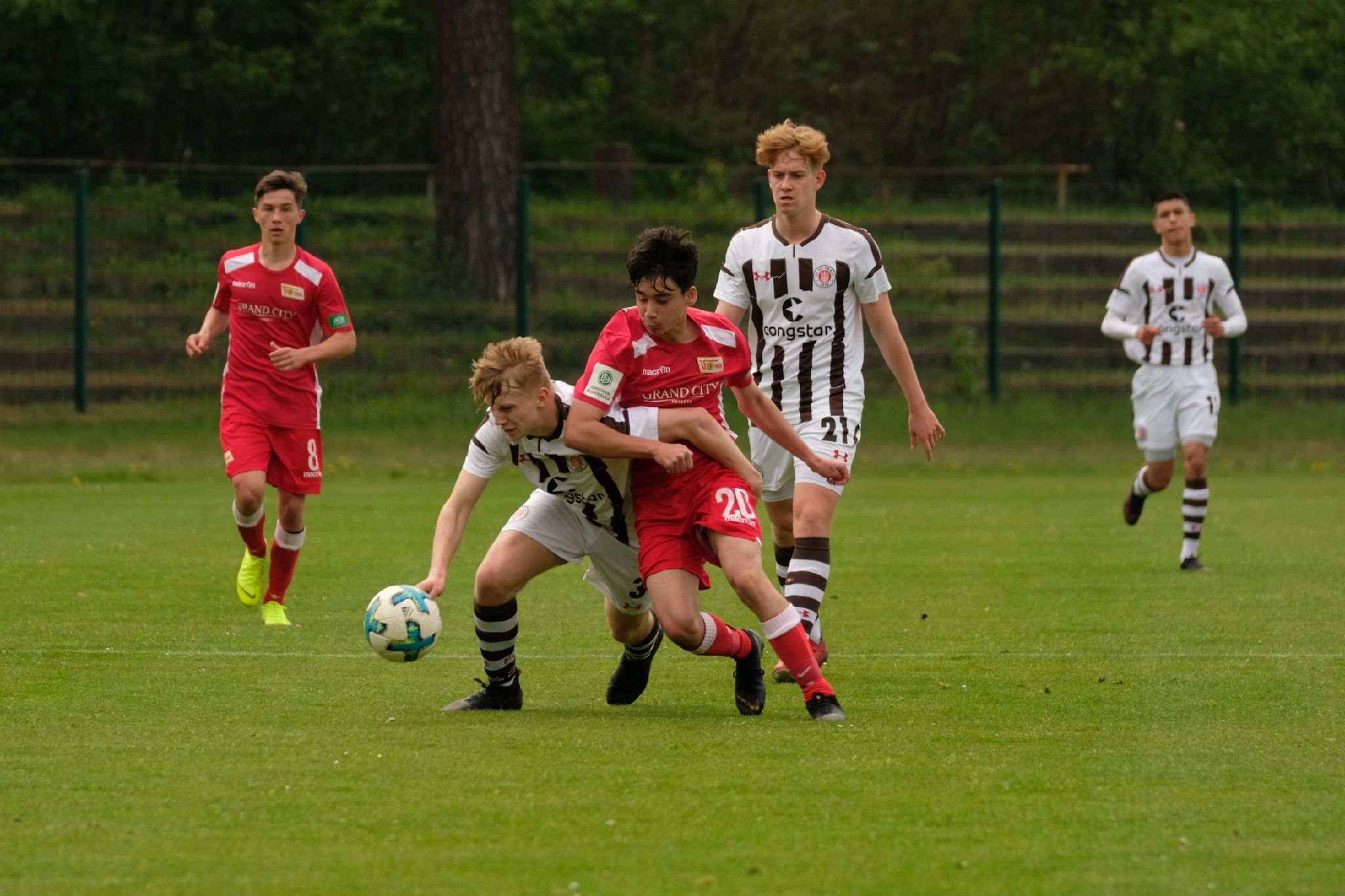 Zwei Fußballspieler im Wettkampf um den Ball auf einem grünen Spielfeld, während andere Spieler in der Nähe stehen.