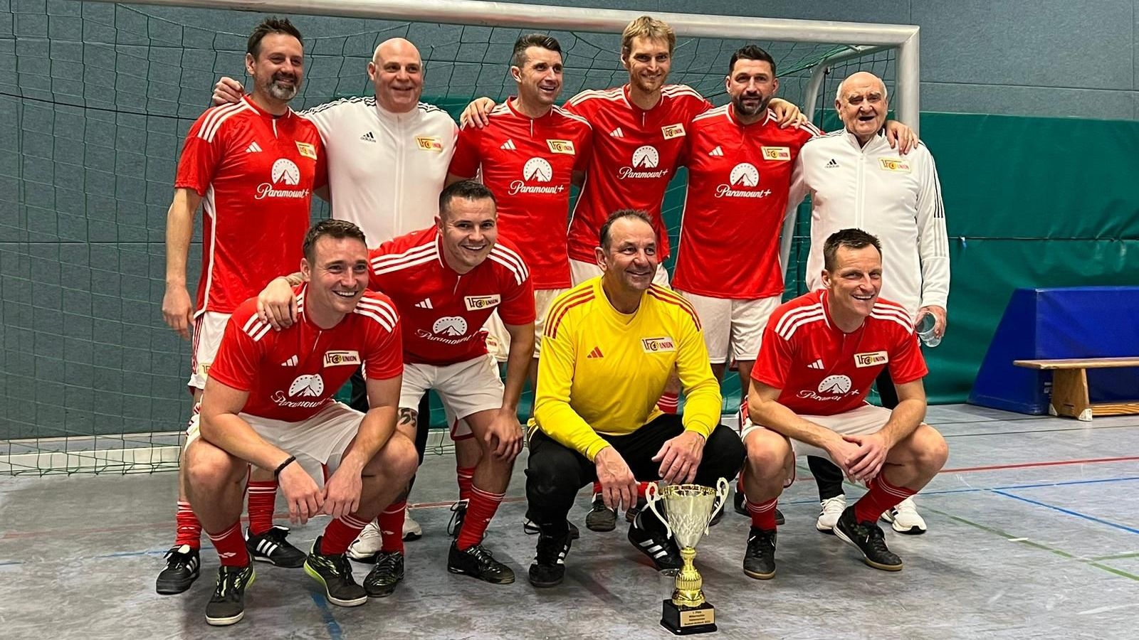Eine Fußballmannschaft in roten Trikots posiert lächelnd mit einem Pokal in der Sporthalle.