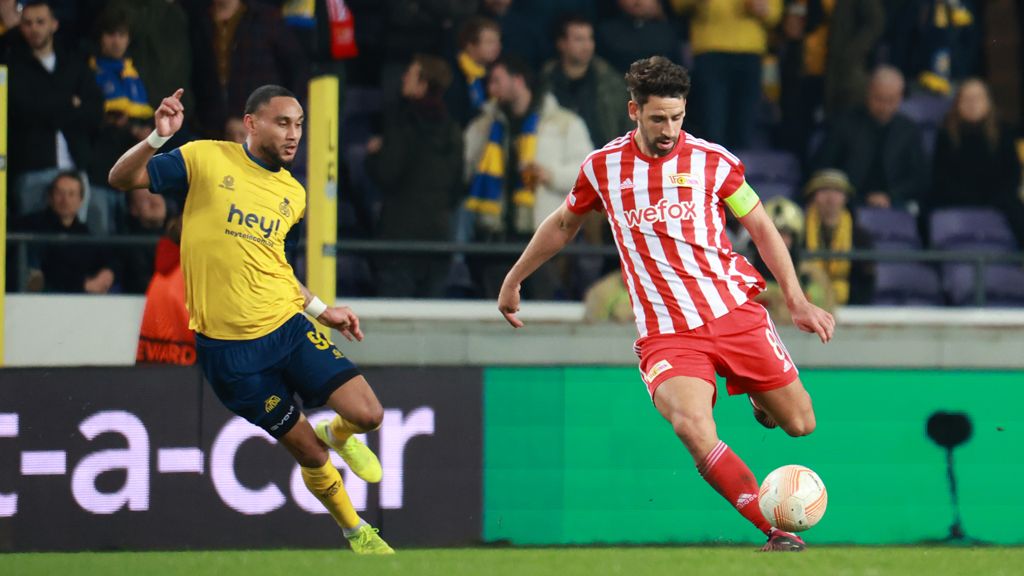 Two soccer players competing for the ball during a game. The player in red and white is dribbling, while the one in yellow is chasing him.