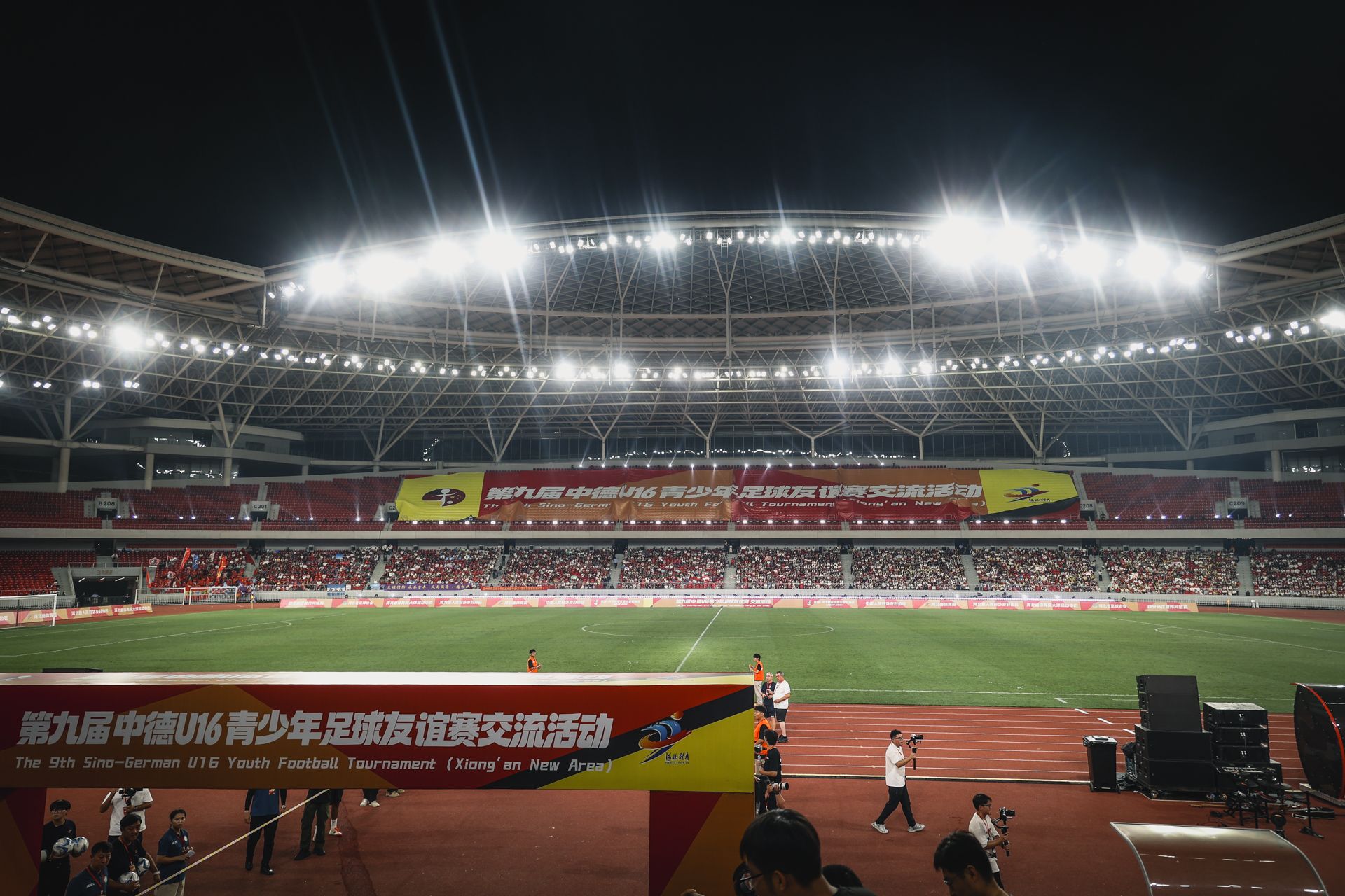 Ein Fußballstadion während eines Jugendturniers, mit Zuschauertribünen und heller Beleuchtung.