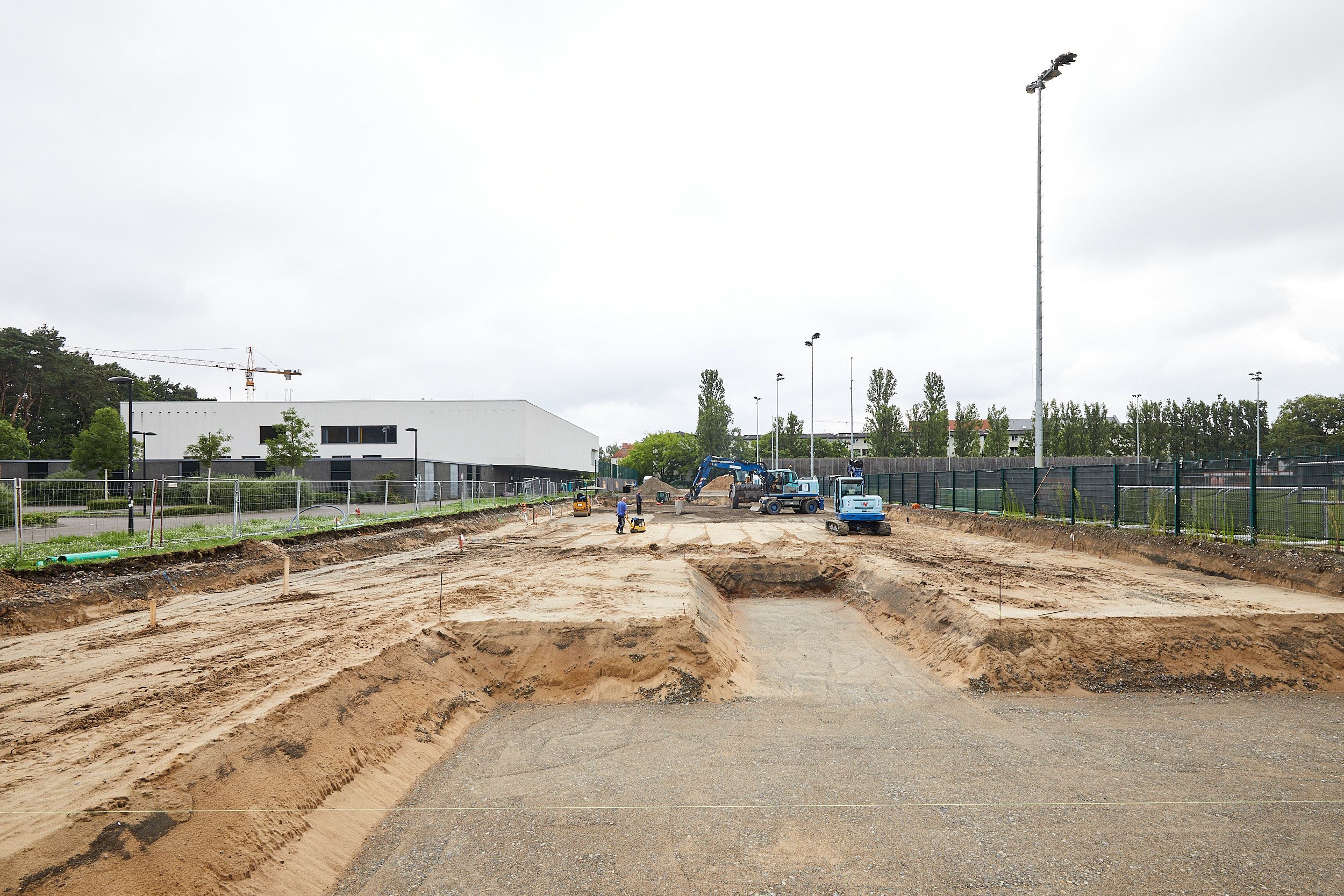 Construction site with earth and sand, where two machines and workers are active, surrounded by a fenced area.