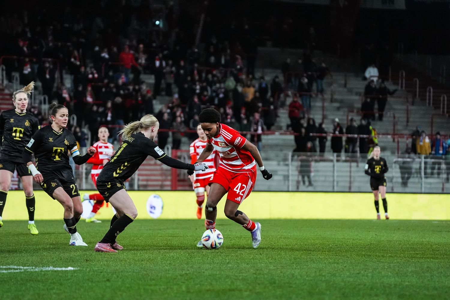 Spielszene im Frauenfußball: Eine Spielerin im roten Trikot dribbelt den Ball, während eine Gegnerin im schwarzen Trikot versucht, ihn zu stoppen.
