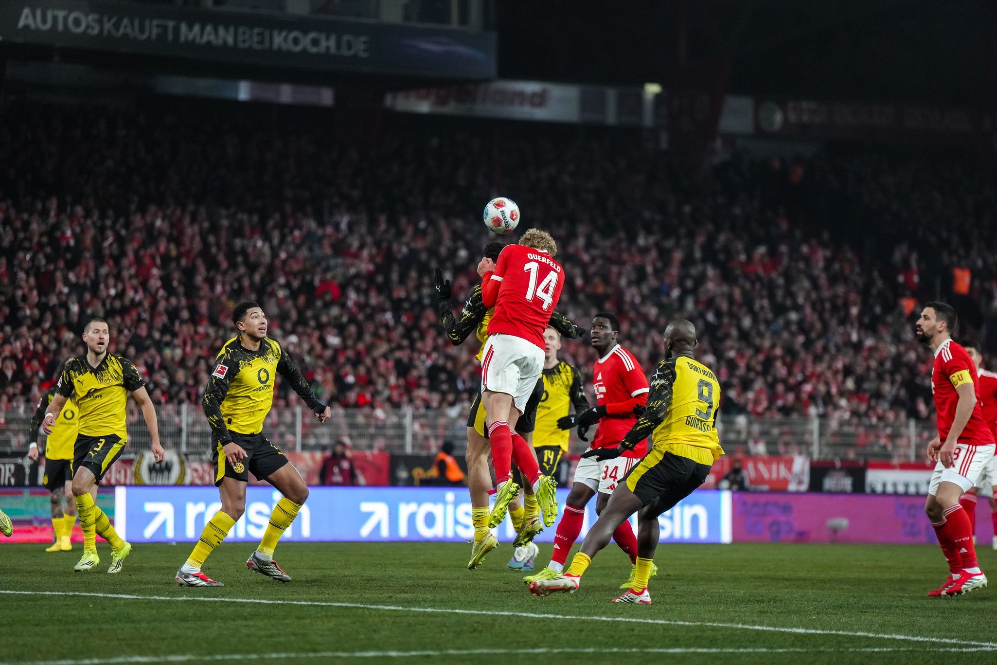 Spielszene im Fußballspiel, Spieler heben ab, um einen Ball hoch in der Luft zu köpfen, mit Spielern und jubelndem Publikum im Hintergrund.