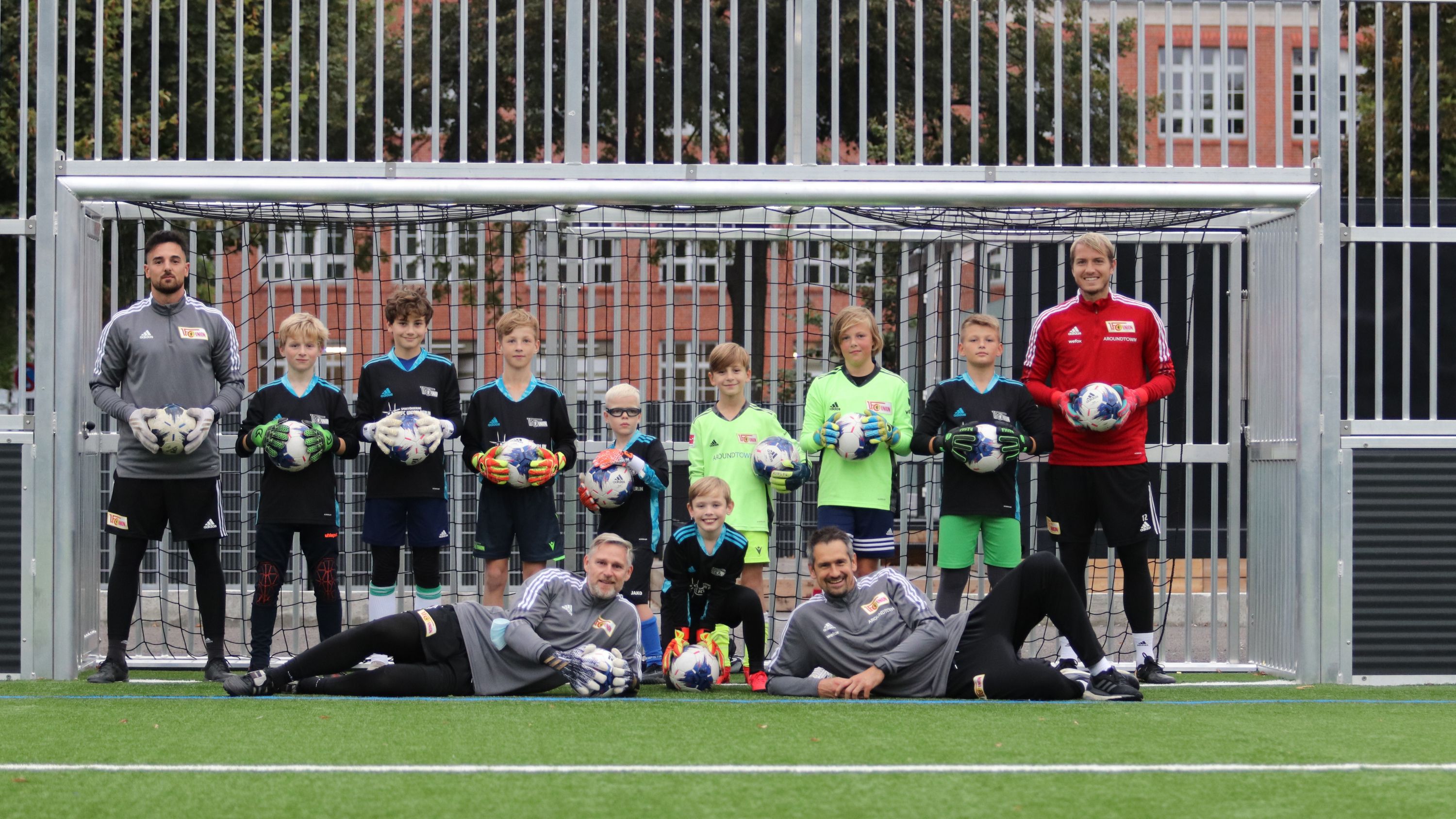 Eine Gruppe von Kindern in Fußballtrikots, die Tore und Bälle halten, steht vor einem Fußballtor mit Trainern.