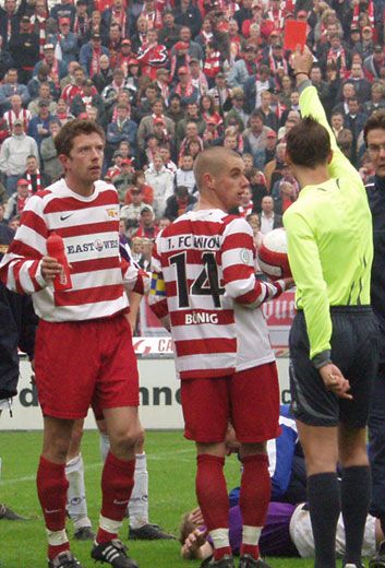 Zwei Fußballspieler in gestreifter rot-weißer Kleidung stehen auf dem Spielfeld. Ein Schiedsrichter zeigt eine rote Karte.