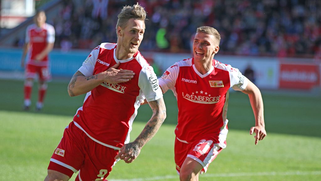 Zwei Fußballspieler in roten Trikots freuen sich. Sie haben ein Tor geschossen. Einer zeigt auf sein Herz. Der andere lacht.