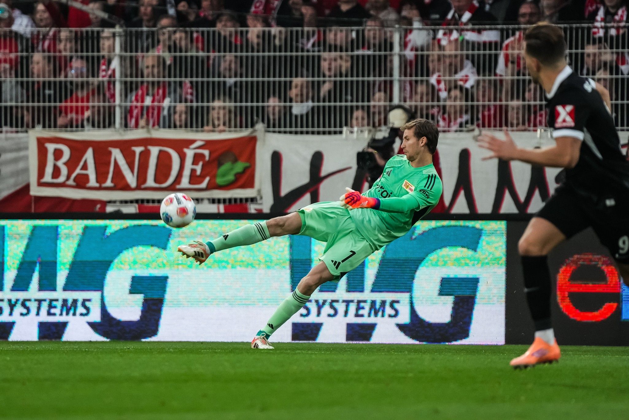 Torwart in grünem Trikot führt einen Schuss mit dem rechten Fuß aus, während ein Spieler in Schwarz bereit ist. Fans im Hintergrund.