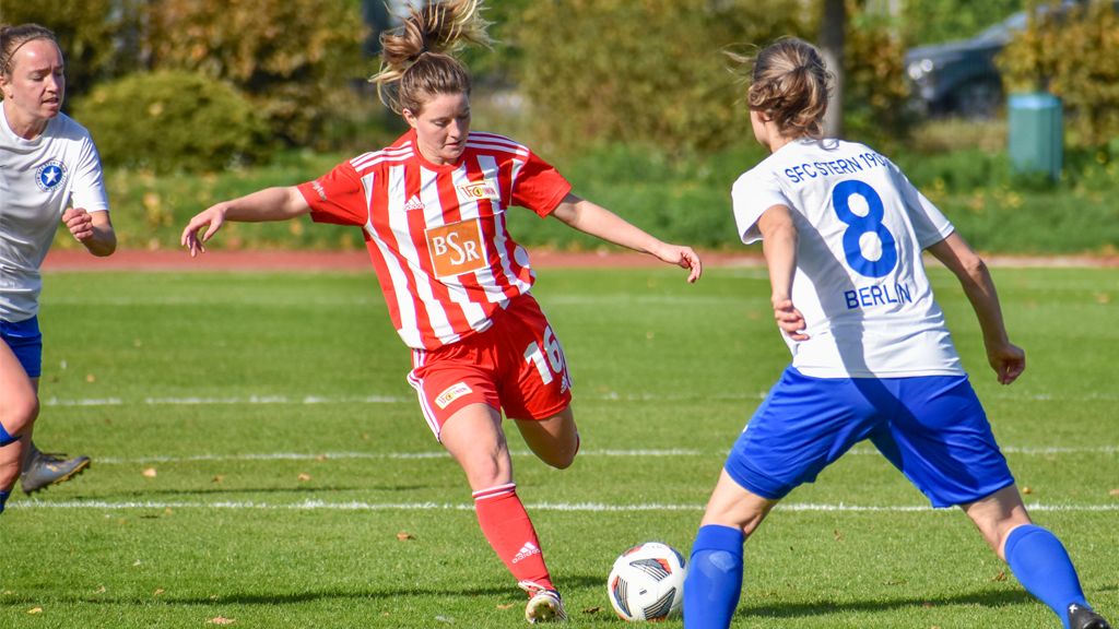 Zwei Fußballspielerinnen auf dem Feld, eine in roter und weißer Uniform, die andere in blauer, während ein Ball zwischen ihnen rollt.