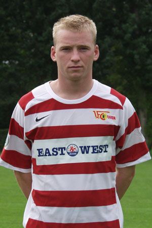 Ein junger Mann in einem gestreiften Fußballtrikot mit dem Logo eines Vereins steht vor einem grünen Hintergrund.