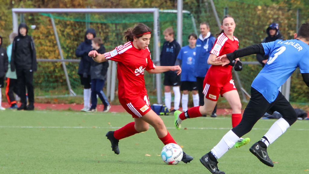 Ein junges Mädchen im roten Fußballtrikot dribbelt den Ball auf einem Rasenplatz, während ein Spieler in Blau angreift. Zuschauer im Hintergrund.