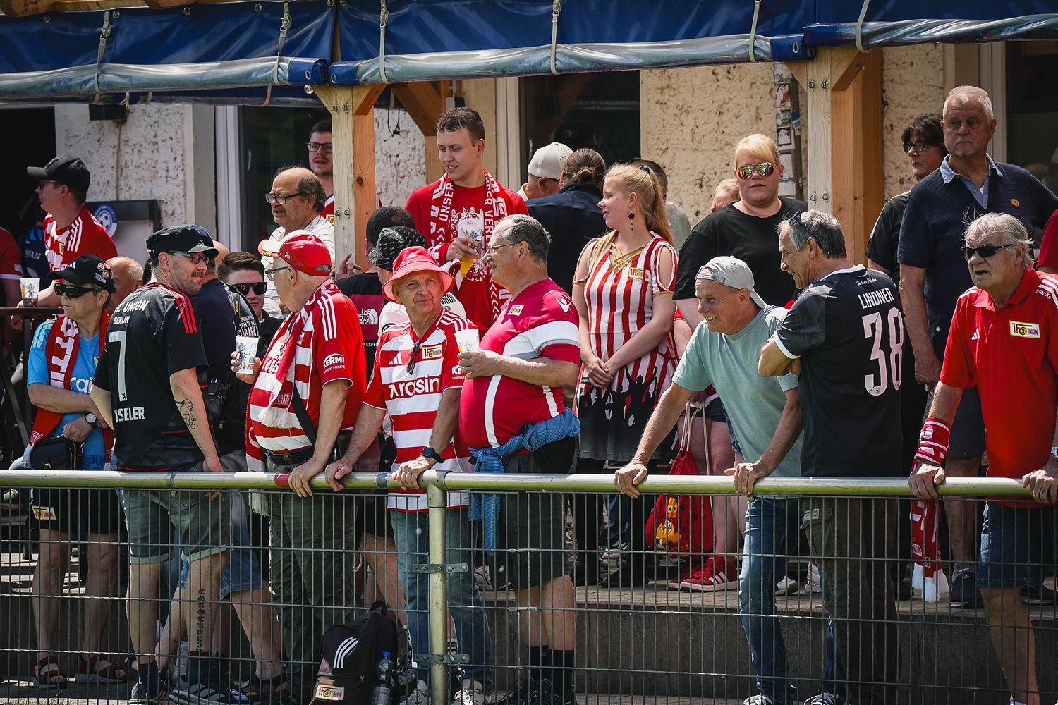 Eine Gruppe von Fans in rot-weißen Trikots steht an einem Zaun, umgeben von weiterer Zuschauerschaft.