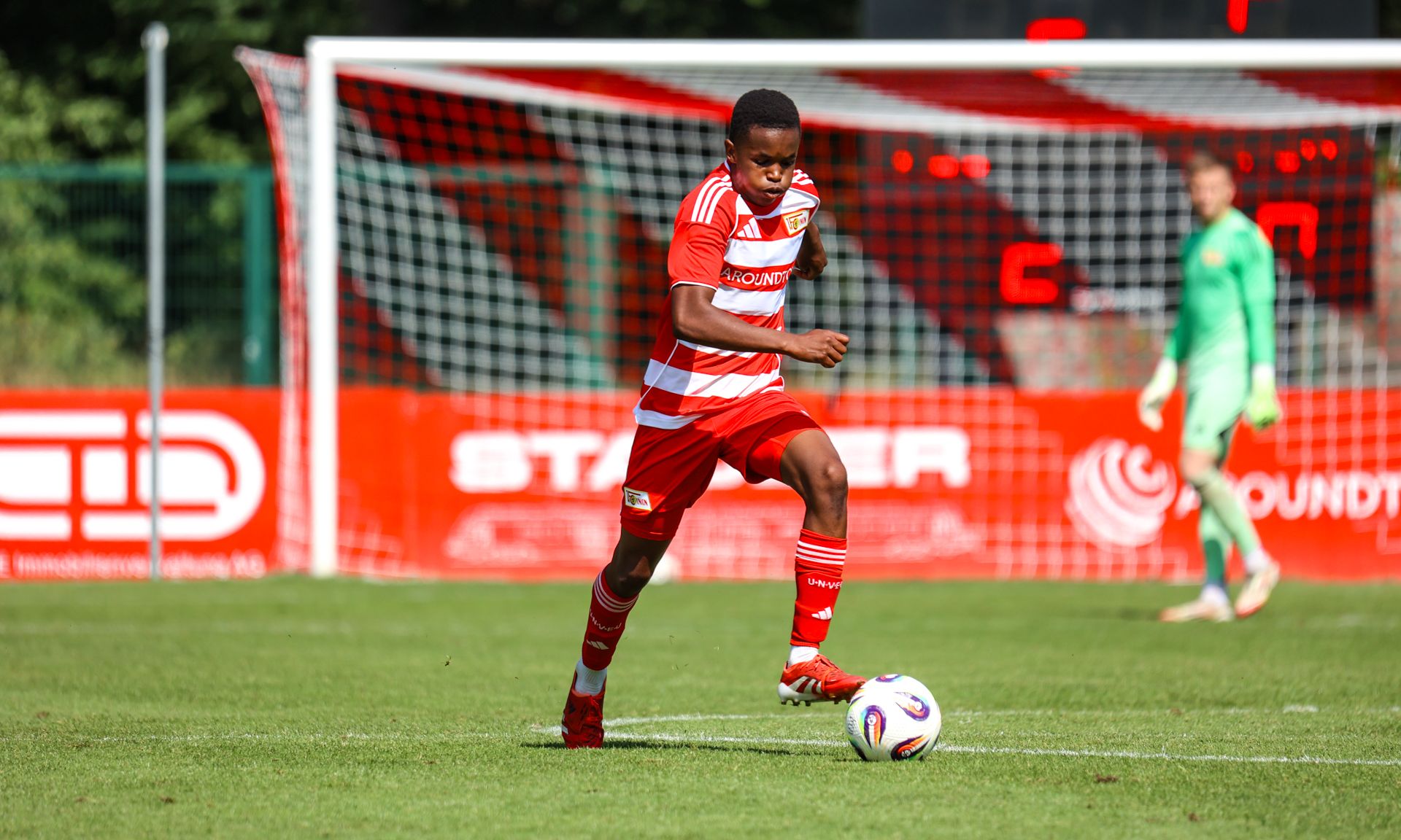 Ein Fußballspieler in rot-weißer Kleidung spielt mit dem Ball. Im Hintergrund steht ein Torwart.