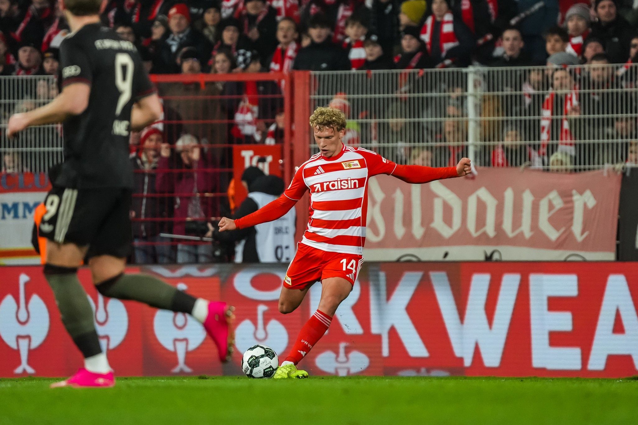 Ein Fußballspieler in rotem Trikot führt einen Freistoß aus, während Zuschauer im Hintergrund jubeln.