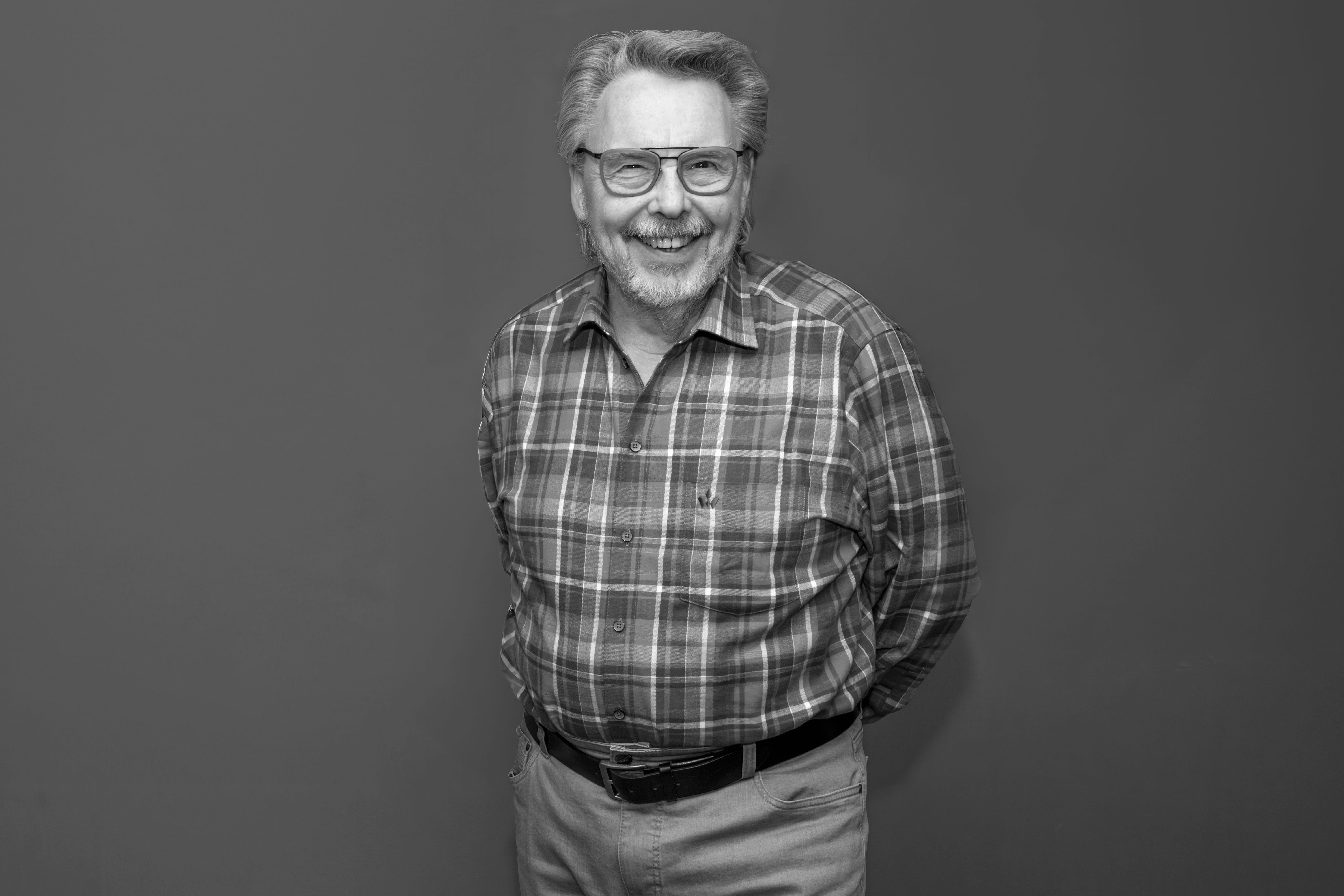 Older man with glasses and an empty smile in a checked shirt, standing in front of a solid color background.