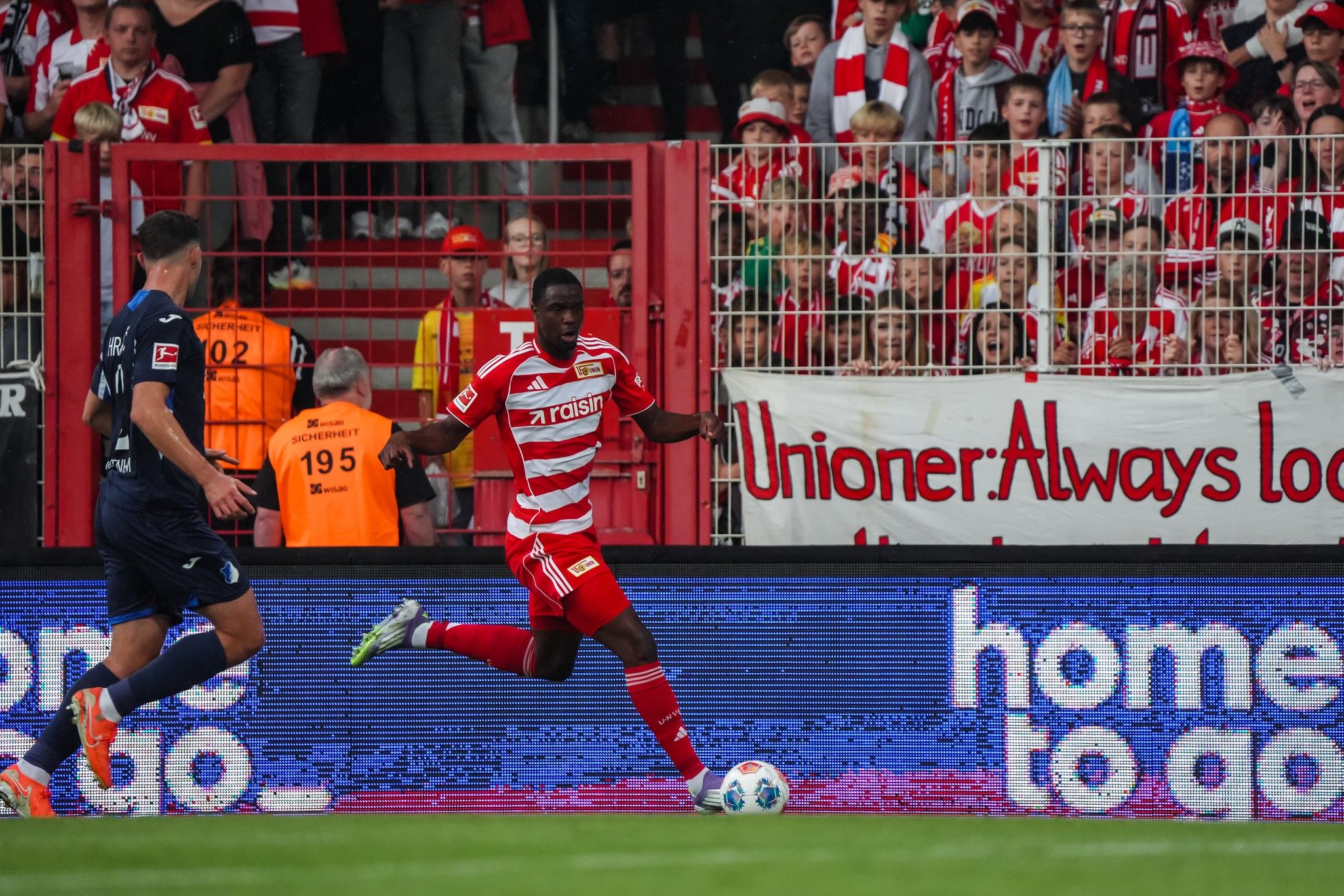 Ein Fußballspieler in rot-weißer Kleidung dribbelt den Ball, während Fans im Hintergrund jubeln.