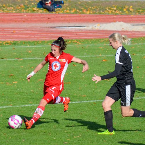 Zwei Fußballspielerinnen im Wettkampf: Eine in rotem Trikot schießt den Ball, die andere in schwarzer Kleidung verfolgt sie.