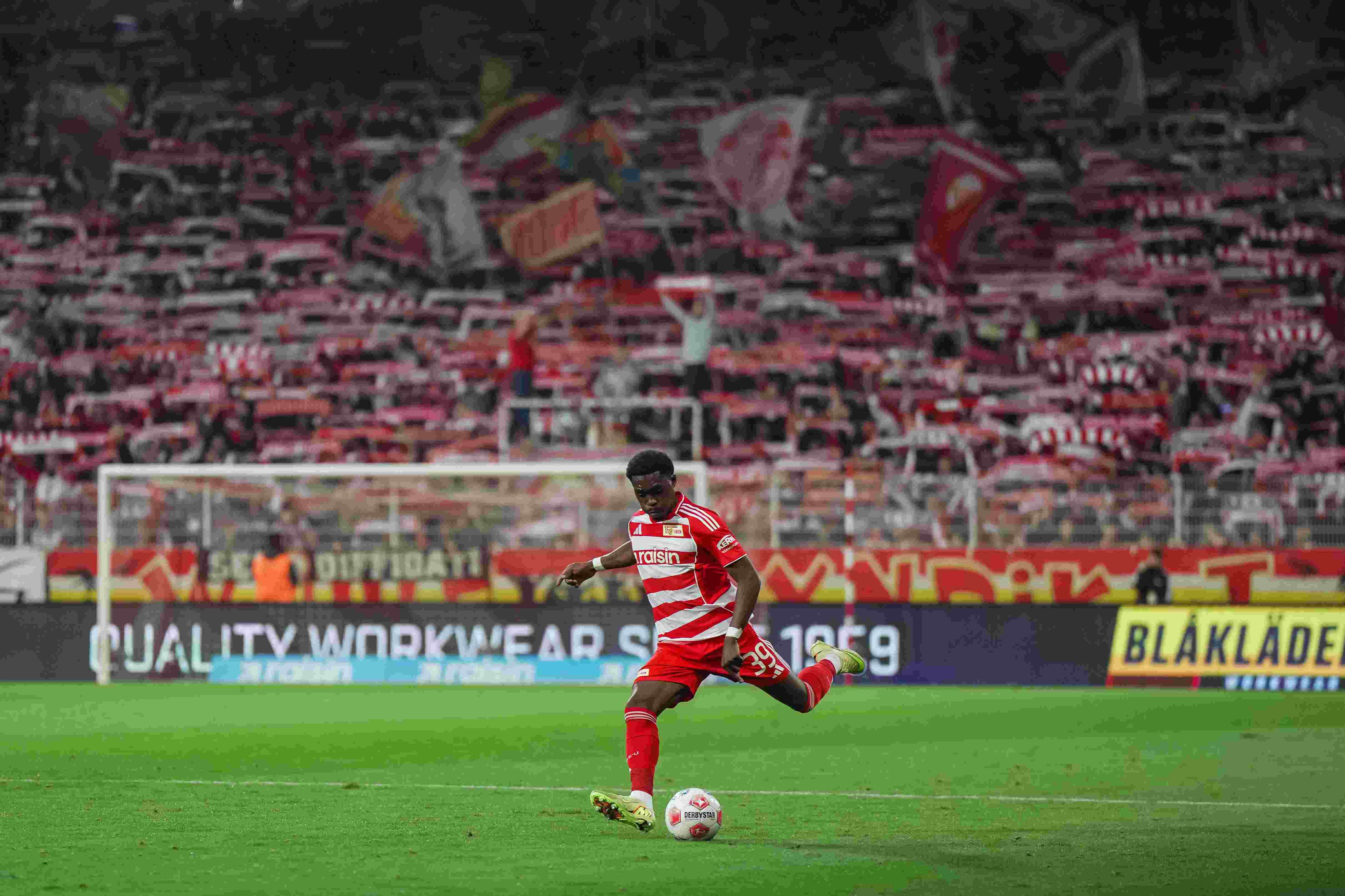 Ein Spieler mit rotem Trikot schießt den Ball. Im Hintergrund jubeln die Fans. Viele rote und weiße Fahnen sind zu sehen.