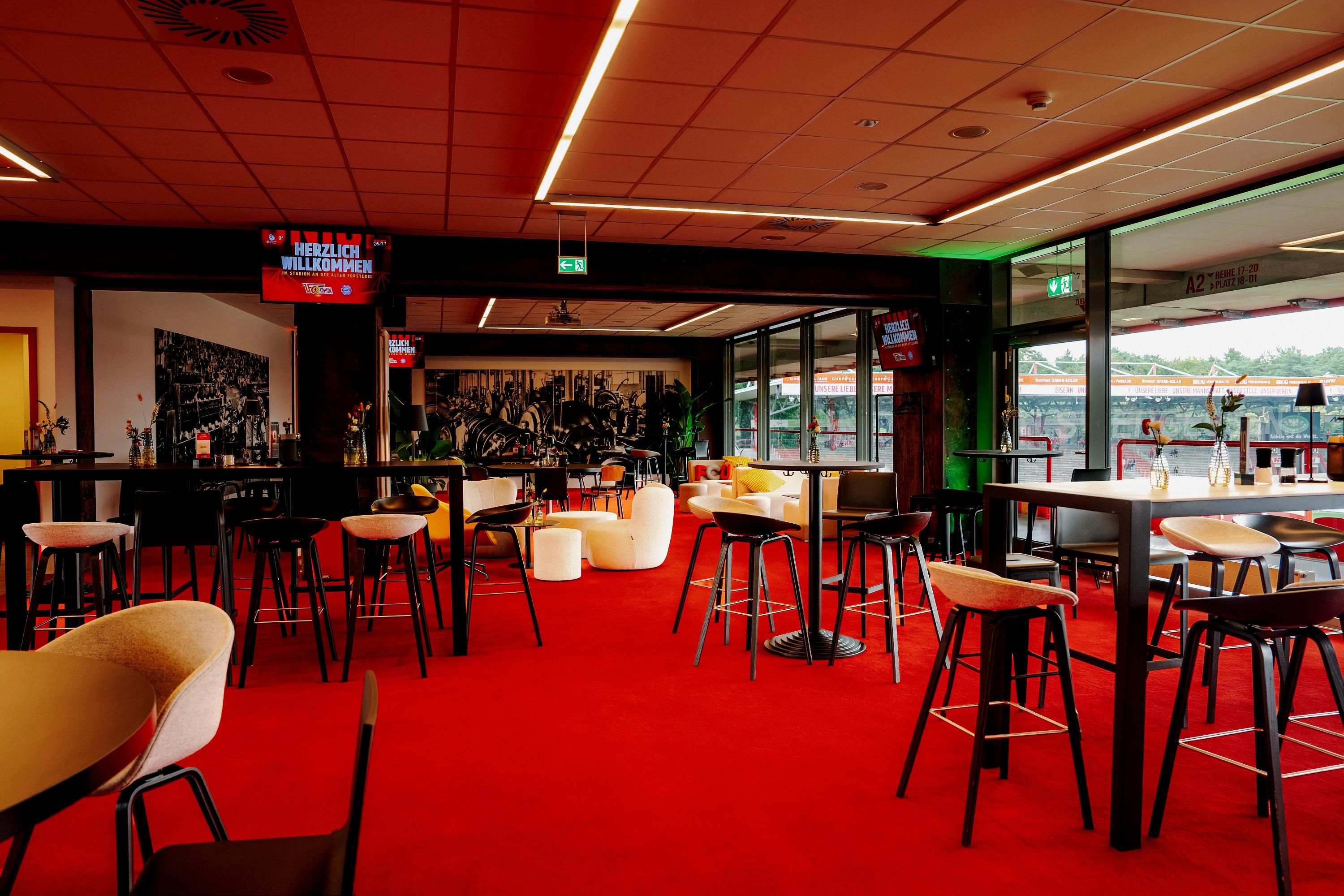 Moderne Bar- und Loungeeinrichtung mit roten Teppichen, hohen Tischen und Stühlen, sowie großen Fenstern und digitaler Anzeige.