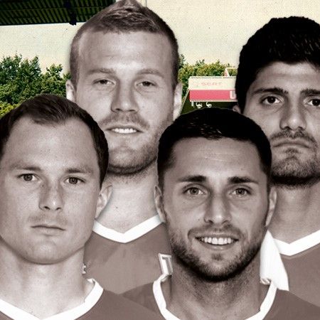 Fünf Fußballspieler in roten Trikots, eng nebeneinander stehend, vor einem unscharfen Stadionhintergrund.