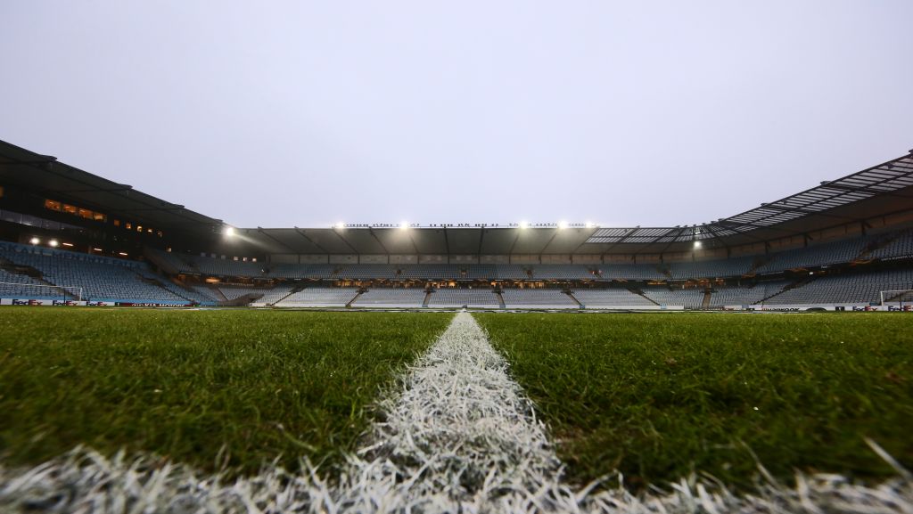 Leeres Fußballstadion bei bewölktem Himmel, mit grünem Rasen und einer weißen Mittellinie im Vordergrund.