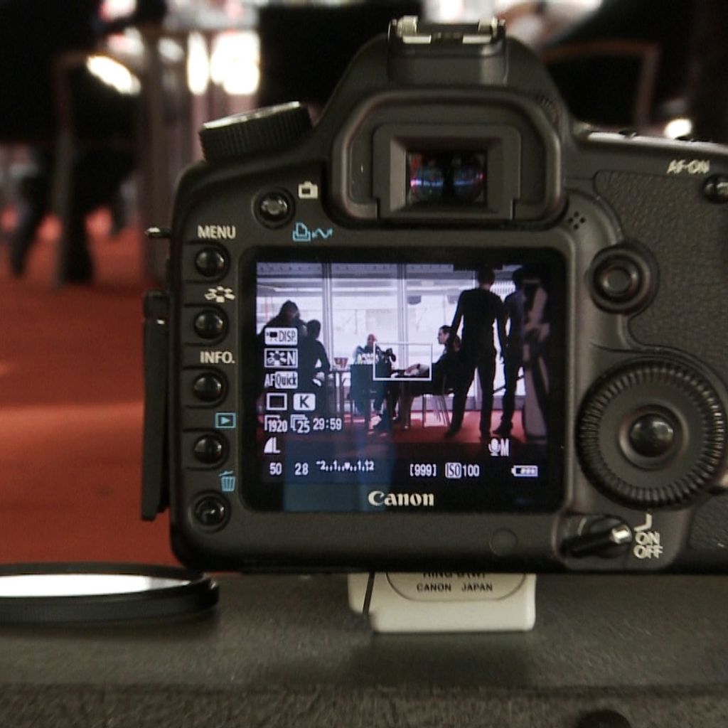 Ein Blick durch die Kamera auf eine Szene in einem hellen Raum mit Personen im Hintergrund.