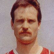 Ein Porträt eines Mannes mit dunklem Haar und einem Schnurrbart, der ein rotes Sporttrikot trägt.