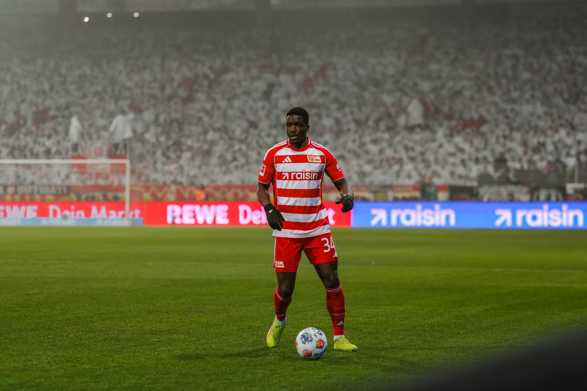 Spieler in rotem Trikot mit weißen Streifen steht auf dem Fußballfeld, bereit für den Spielzug, im Hintergrund jubelnde Fans.