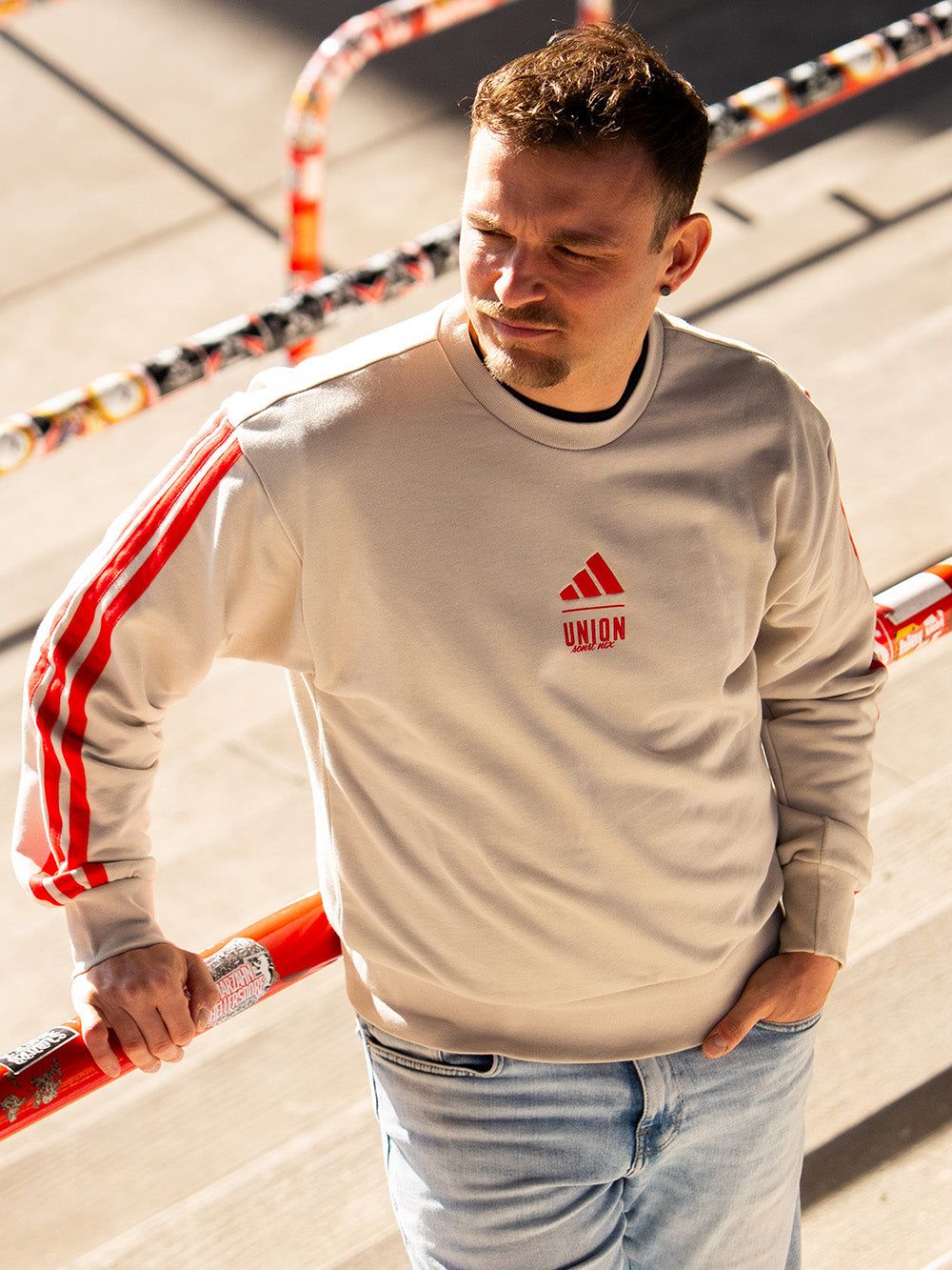 Ein Mann mit kurzem Haar trägt einen beigen Sweatshirt mit roten Streifen und steht an einer abgeschrägten Fläche.