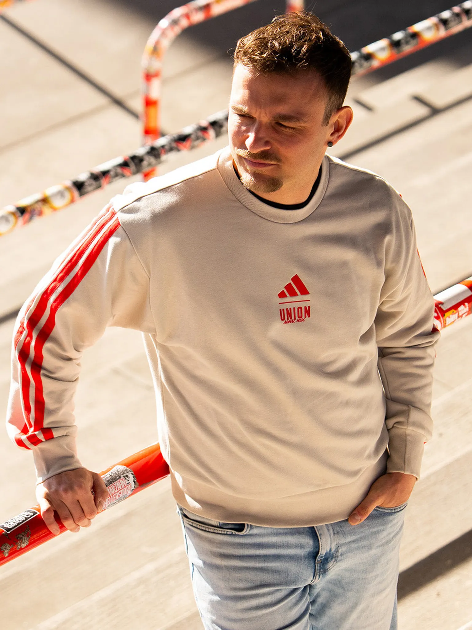 Ein Mann mit kurzem Haar trägt einen beigen Sweatshirt mit roten Streifen und steht an einer abgeschrägten Fläche.