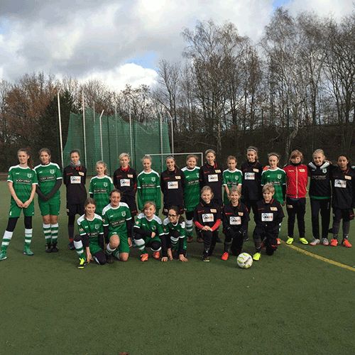 Mädchen-Fußballmannschaft posiert auf einem Platz, einige tragen grüne und schwarze Trikots, im Hintergrund sind Bäume und ein Tor.