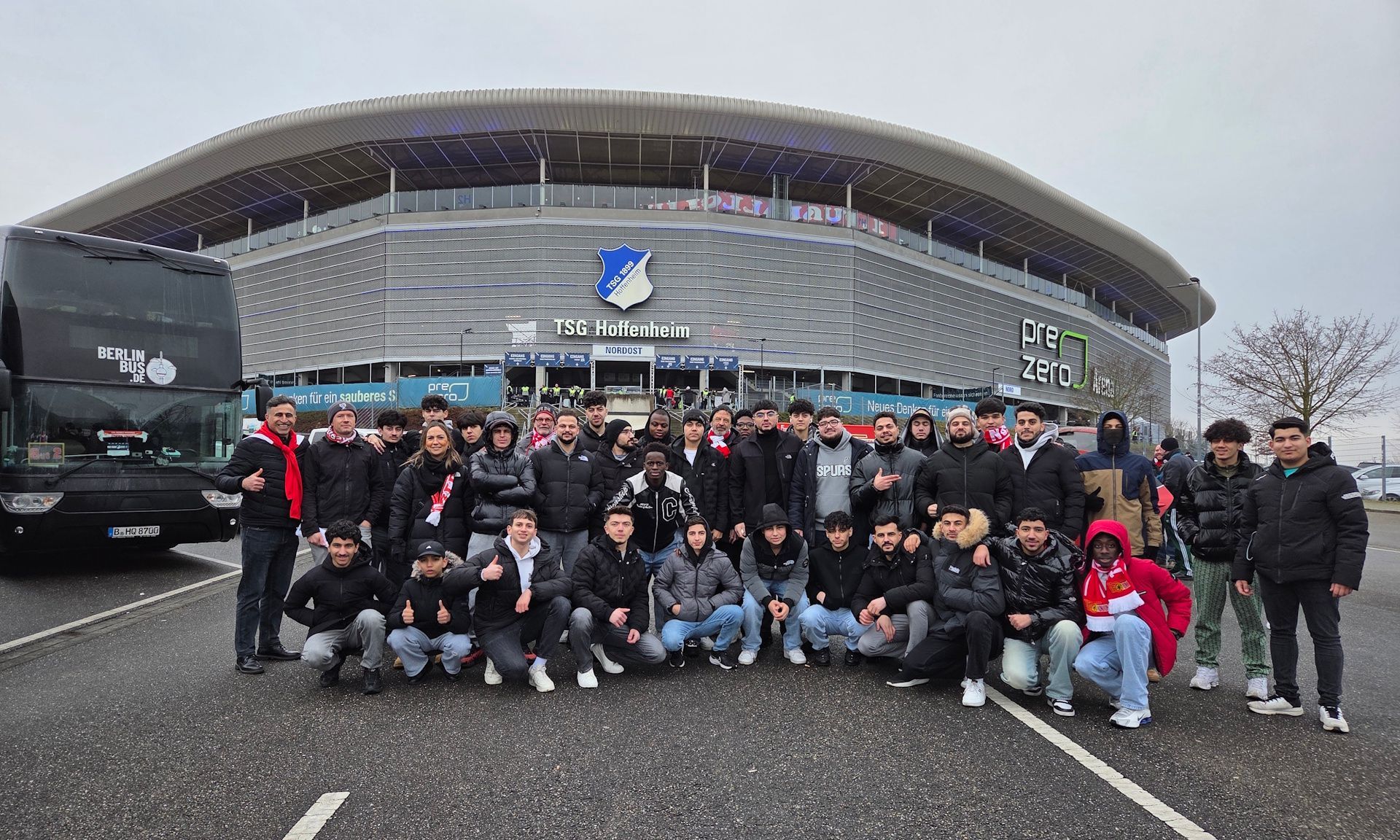 Gruppe von Menschen in einheitlicher Kleidung vor dem Stadion der TSG Hoffenheim, umgeben von Bussen und einer wolkenverhangenen Himmel.