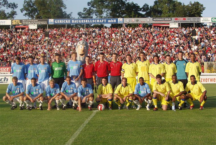 Zwei Fußballmannschaften posieren gemeinsam auf einem Rasenfeld vor einer großen Zuschauermenge im Hintergrund.