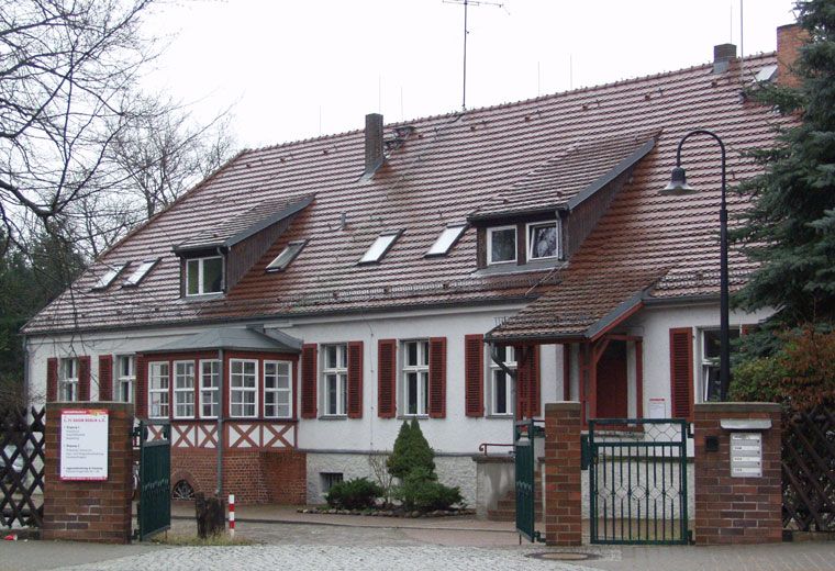 Historisches Gebäude mit rotem Ziegeldach, Fensterläden und einem Gartenzaun, umgeben von Bäumen und Sträuchern.