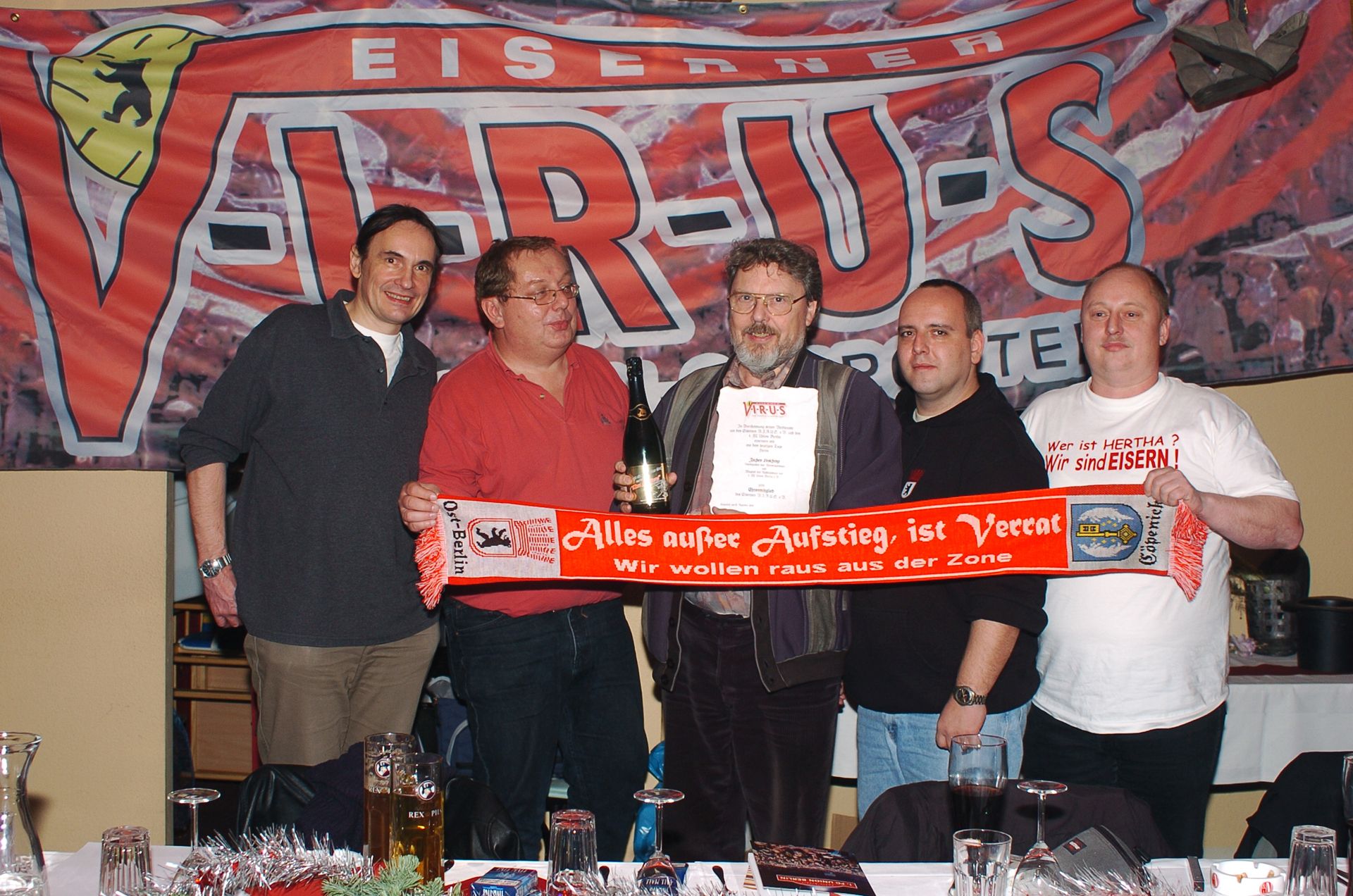 Eine Gruppe von fünf Männern steht lächelnd vor einem Banner mit der Aufschrift "VIRUS" und hält ein Zertifikat sowie einen Schal.