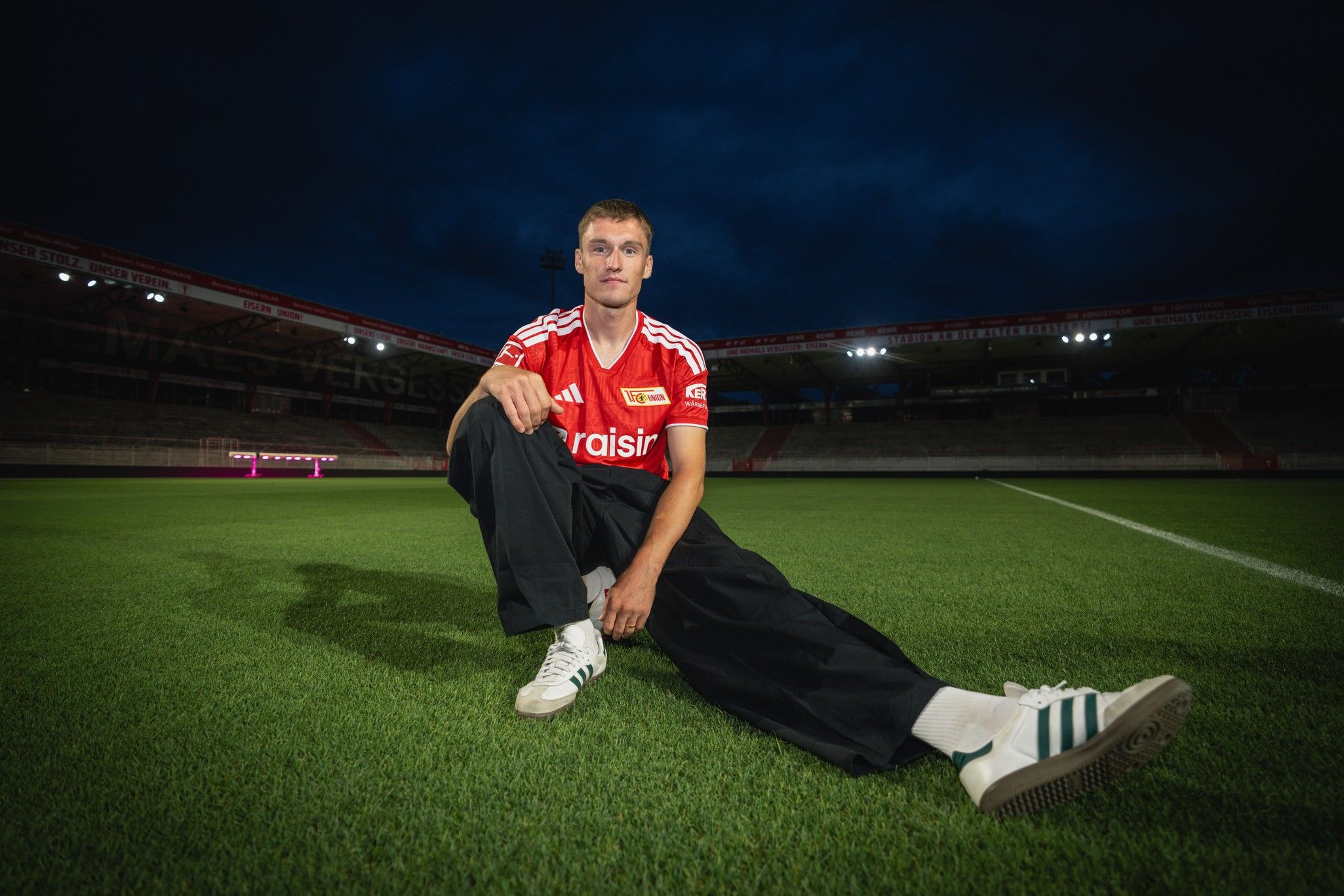 Ein junger Mann sitzt auf dem Fußballfeld in einem Stadion, trägt ein rotes Trikot und schwarze Hose, mit einem düsteren Himmel im Hintergrund.