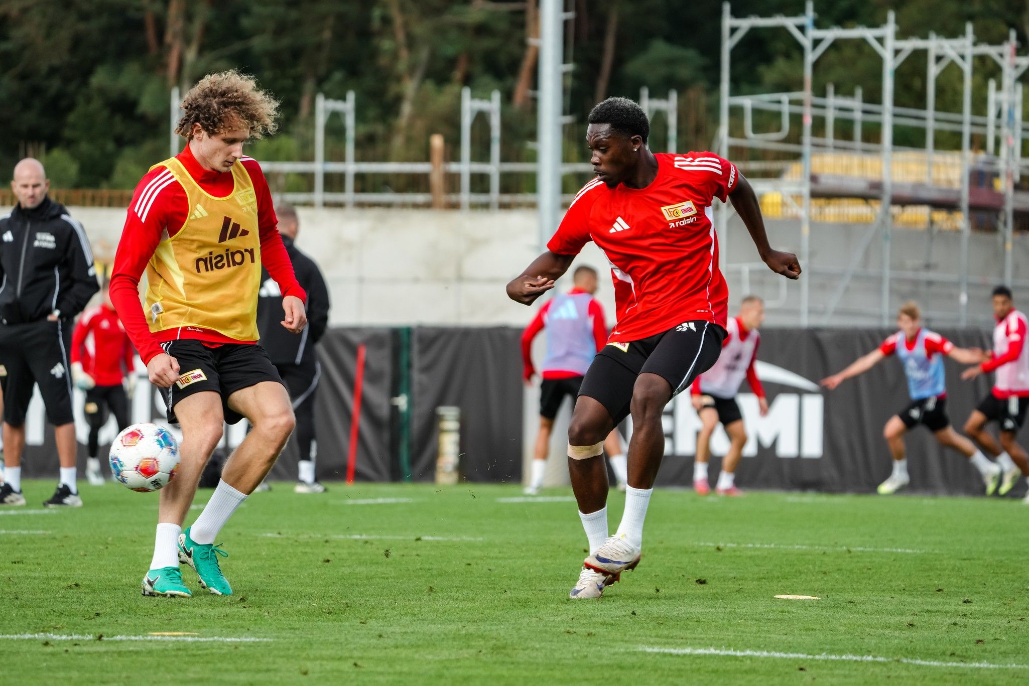 Zwei Fußballspieler in Trainingskleidung üben auf dem Platz, während weitere Spieler im Hintergrund trainieren.