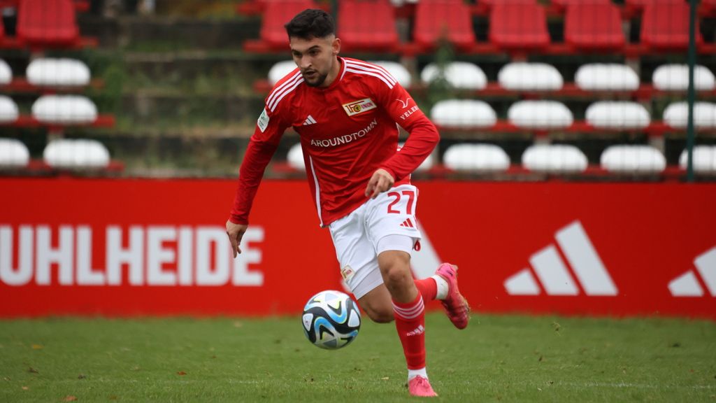 Ein Fußballspieler in rotem Trikot mit der Nummer 27 dribbelt über das Spielfeld mit einer grünen Rasenfläche im Hintergrund.