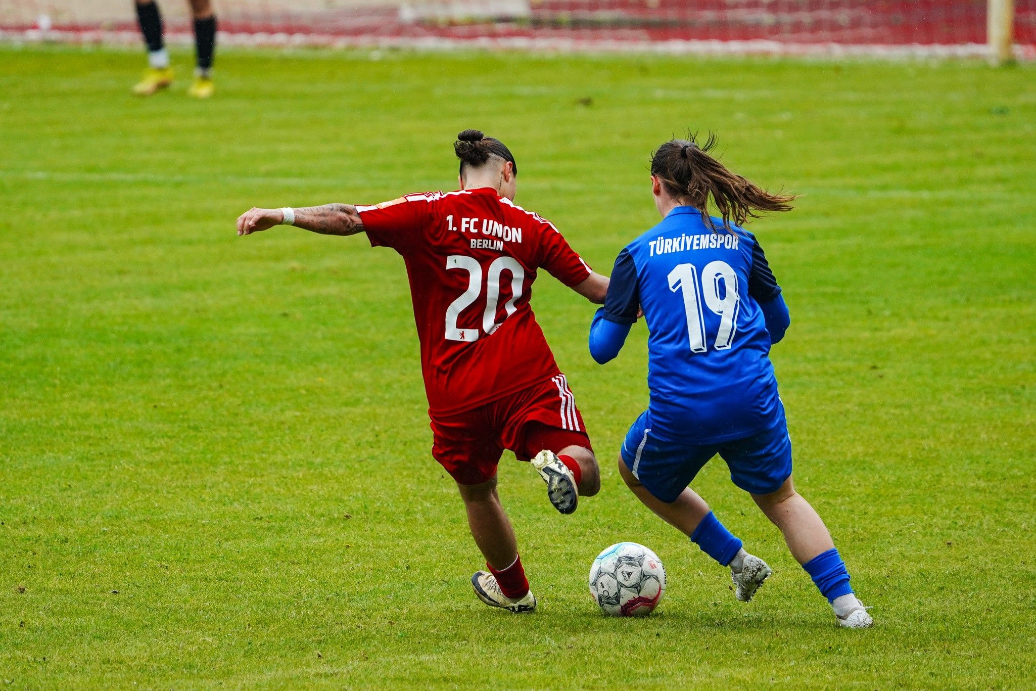 Spielszene eines Fußballspiels: Eine Spielerin in rotem Trikot dribbelt, während eine andere in blauem Trikot sie verfolgt.