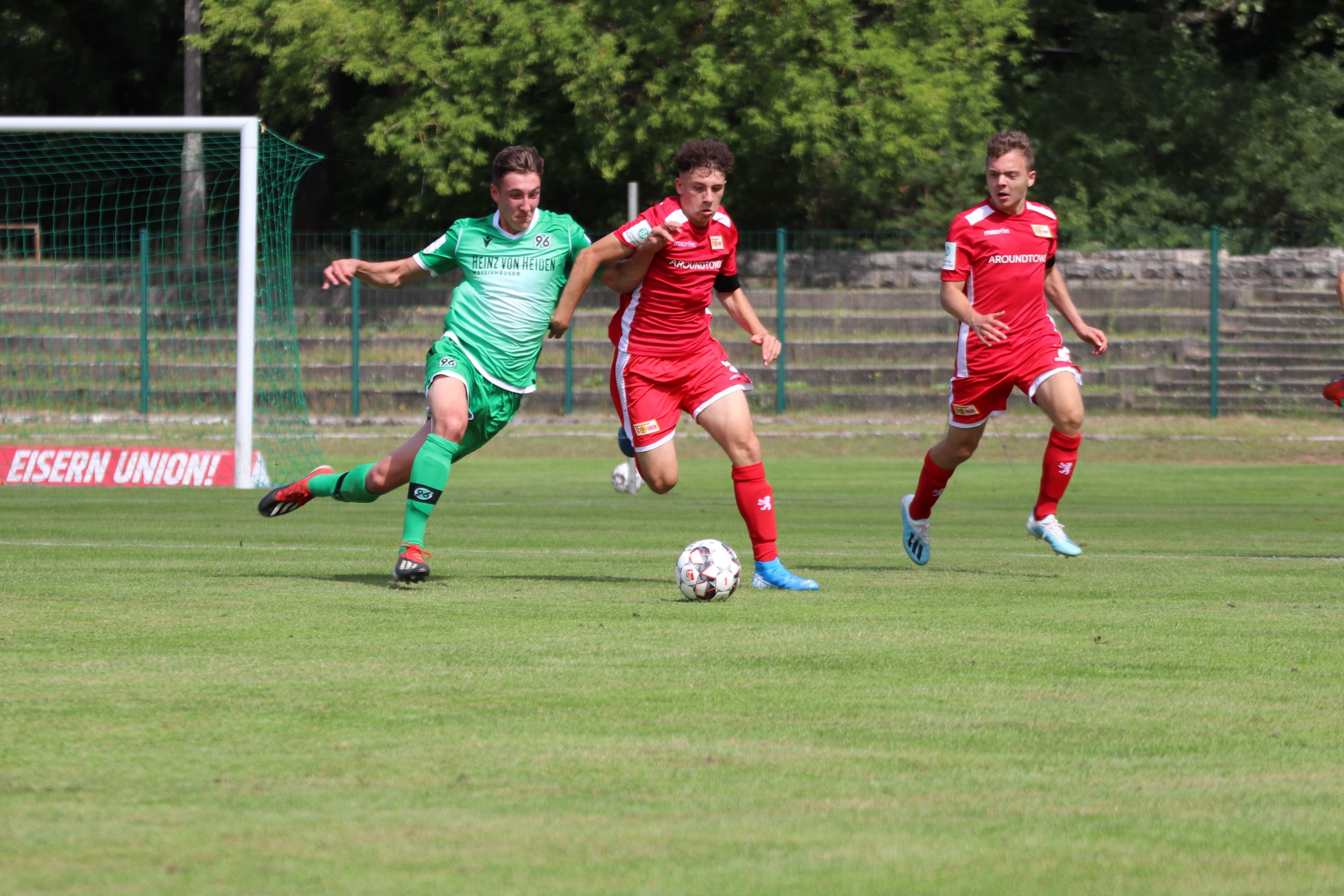 Ein Fußballspiel. Ein Spieler trägt ein grünes Trikot. Er dribbelt den Ball. Zwei Gegner laufen hinter ihm her.