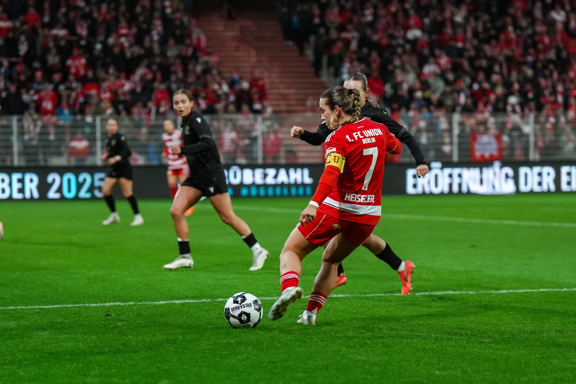 Spielszene im Fußball mit Spielerin in rotem Trikot, die den Ball an einer Gegnerin vorbeispielt. Hintergrund mit Zuschauern im Stadion.